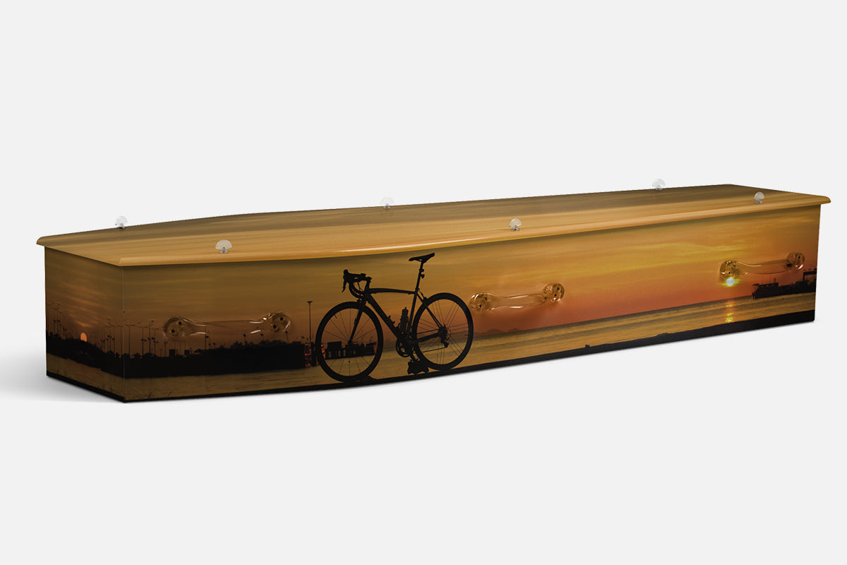 Cycling coffin with a silhouetted road bike leaning at a waterside sunset with a ship on the horizon