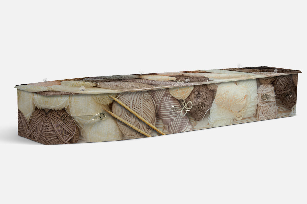 Knitting coffin in a photographic pile of cream and brown wool balls with knitting needles resting among them